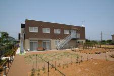 新築の介護施設の外観。赤茶色のレンガ造り2階建てで、前庭には新しく植えられた樹木が見られます。